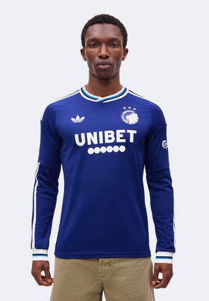 Homme portant un maillot de football bleu à manches longues du FC Copenhague avec des logos blancs et un pantalon beige, debout devant un fond uni.