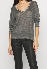 Szary, luźny top z długim rękawem i głębokim dekoltem w kształcie litery V, z podwiniętymi mankietami i subtelną fakturą. Połączony z czarnymi dopasowanymi spodniami.