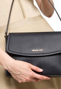 Mano che tiene una borsa a spalla Michael Kors in pelle nera testurizzata con logo dorato, indossando un vestito dorato chiaro con zip visibile.