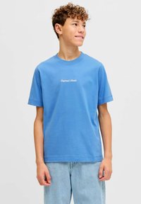 Εφηβικός αγόρι με σγουρά μαλλιά που φοράει μπλε μπλουζάκι με το κείμενο "Originals Studio" και ανοιχτόχρωμα τζιν, κοιτάζοντας προς τα δεξιά του σε ένα απλό φόντο.