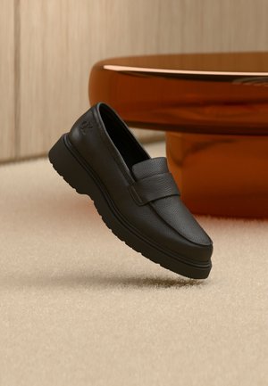 Čierny kožený mokasín s hrubou podrážkou na béžovom koberci, v pozadí lesklý hnedý okrúhly kus nábytku.