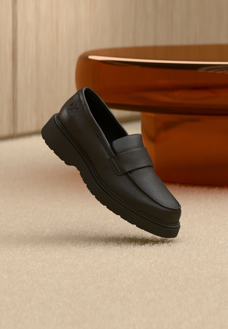 Μαύρο δερμάτινο μοκασίνι με παχιά σόλα που ξεκουράζεται σε μπεζ χαλί, με ένα γυαλιστερό καφέ κυκλικό έπιπλο στο φόντο.