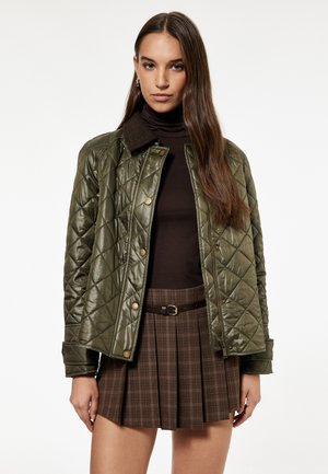 Grüne gesteppte Jacke mit einem braunen Kragen, die goldene Knöpfe hat. Sie wird über einem dunklen Oberteil und einem karierten braunen Faltenrock getragen, der die gestuften Texturen hervorhebt.