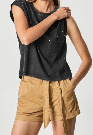 Kobieta w czarnej, bezrękawnikowej bluzce w drobne białe kropki oraz w jasnobrązowych, wysokim stanie szortach z materiałowym paskiem, z ręką w kieszeni, z ręką dotykającą ramienia.