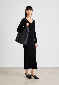 Black fitted long-sleeve dress with a v-neck and a midi hem. Model holds a large black tote bag with a gold accent. Black slingback shoes.