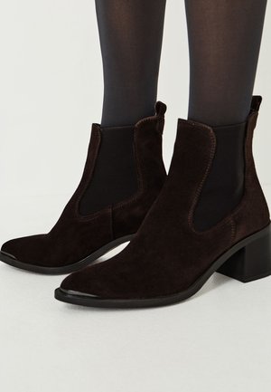Bottines en daim marron avec un talon épais, des panneaux latéraux élastiques et un design à bout pointu. Texture lisse avec des détails de couture contrastants.
