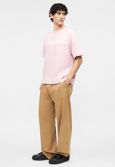 Νεαρός άνδρας όρθιος με τα χέρια στις τσέπες, φοράει φαρδιά ροζ μπλούζα και φαρδιά καφέ παντελόνια με μαύρα αθλητικά παπούτσια, κοιτάζει πλάγια.