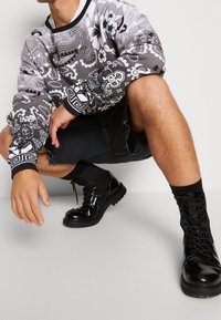 Černá a šedá mikina s květinovými a geometrickými vzory, kombinovaná s potrhanými černými šortkami a lesklými černými vojenskými botami.