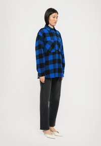 Camicia di flanella a quadri blu e nera oversize con due tasche frontali, abbinata a jeans neri a gamba dritta e mule bianchi.