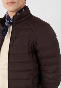 Bruin pufferjack met horizontale stiksels, hoge kraag, ritssluiting en logo-embleem. Draagt dit over een gestructureerde crèmekleurige trui en een marineblauwe sjaal.