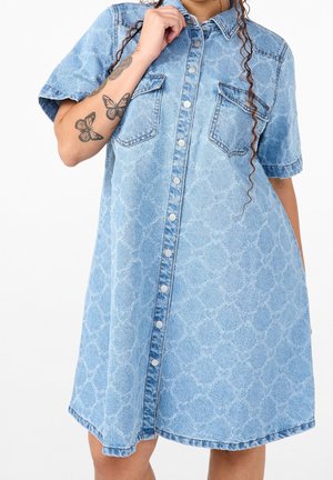 Vrouw draagt een lichtblauwe acid-wash denimjurk met korte mouwen, knopen aan de voorkant, borstzakken en vlindertatoeages op de linker onderarm.