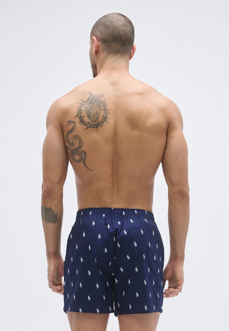 Navy swim trunks with white horse logo pattern, elastic waistband, and a relaxed fit. Features tattoos on a bare back.