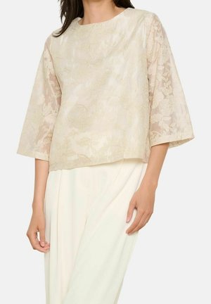 Frau trägt ein elfenbeinfarbenes florales Jacquard-Oberteil mit dreiviertellangen transparenten Ärmeln aus Spitze und weiße, locker sitzende Hosen.