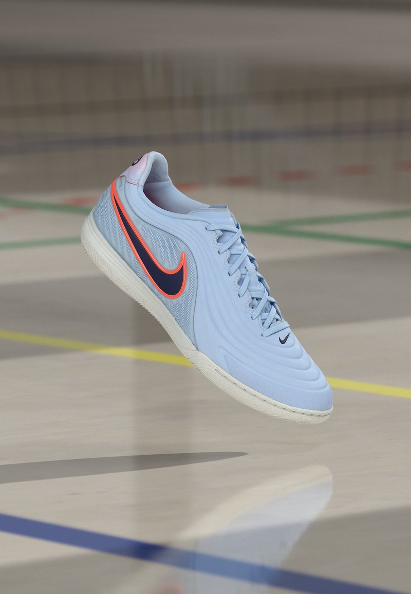 Jasnoniebieski halowy but piłkarski Nike z czarnym logo z pomarańczowymi akcentami, unoszący się nad podłogą sali gimnastycznej z kolorowymi liniami.