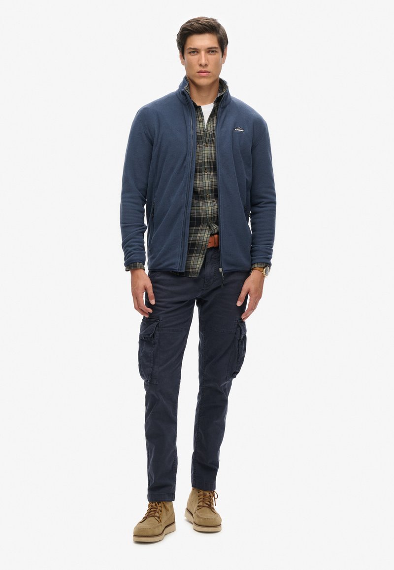 Granatowa polarowa kurtka z zamkiem, koszula w kratkę, spodnie cargo i tanowe buty. Swobodne warstwy z funkcjonalnymi kieszeniami i teksturowanymi materiałami.