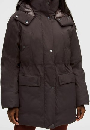 Bruine gewatteerde winterjas met grote knoopsluitingen en hoge kraag, gedragen door een persoon met ontspannen handen langs het lichaam.