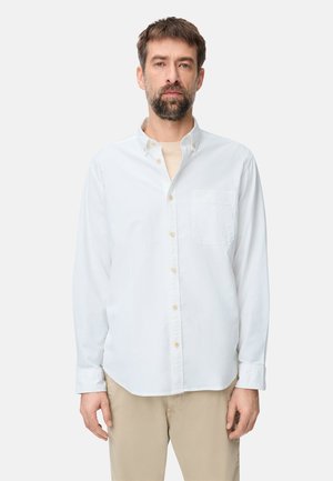 Man met bruin haar en baard, gekleed in een wit overhemd over een beige shirt en beige broek, tegen een effen lichte achtergrond.