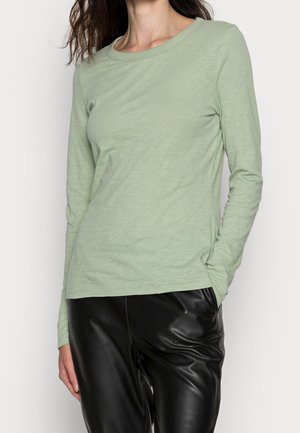 Vrouw die een lichtgroen shirt met lange mouwen en zwarte leren broek draagt, staand met één hand in de zak tegen een effen achtergrond.