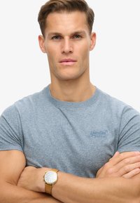 T-shirt gris à manches courtes avec un col rond et un petit logo bleu sur la poitrine. Montre avec bracelet en cuir jaune-brun au poignet.