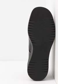 Semelle d’une chaussure noire avec motifs texturés antidérapants, positionnée verticalement contre un angle blanc de mur et de sol.