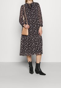 Robe midi noire à motif floral avec manches longues transparentes, col à nouer et taille smockée. Associée à un petit sac rose texturé et des bottines noires.