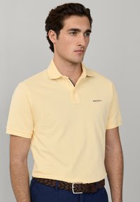 Jeune homme portant un polo à manches courtes jaune clair et un pantalon bleu foncé avec une ceinture tressée marron, debout devant un fond gris uni.
