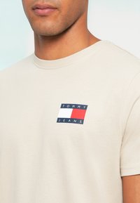 Μπεζ βαμβακερή μπλούζα T-shirt με στρογγυλή λαιμόκοψη. Διαθέτει μια μικρή, χρωματιστή ετικέτα λογότυπου με ναυτικό μπλε, λευκά και κόκκινα ορθογώνια. Κοντά μανίκια.