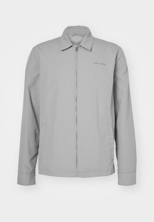 Chaqueta ligera gris con cuello, cremallera frontal, bolsillos laterales y un sutil logotipo en el lado izquierdo del pecho. Textura suave.