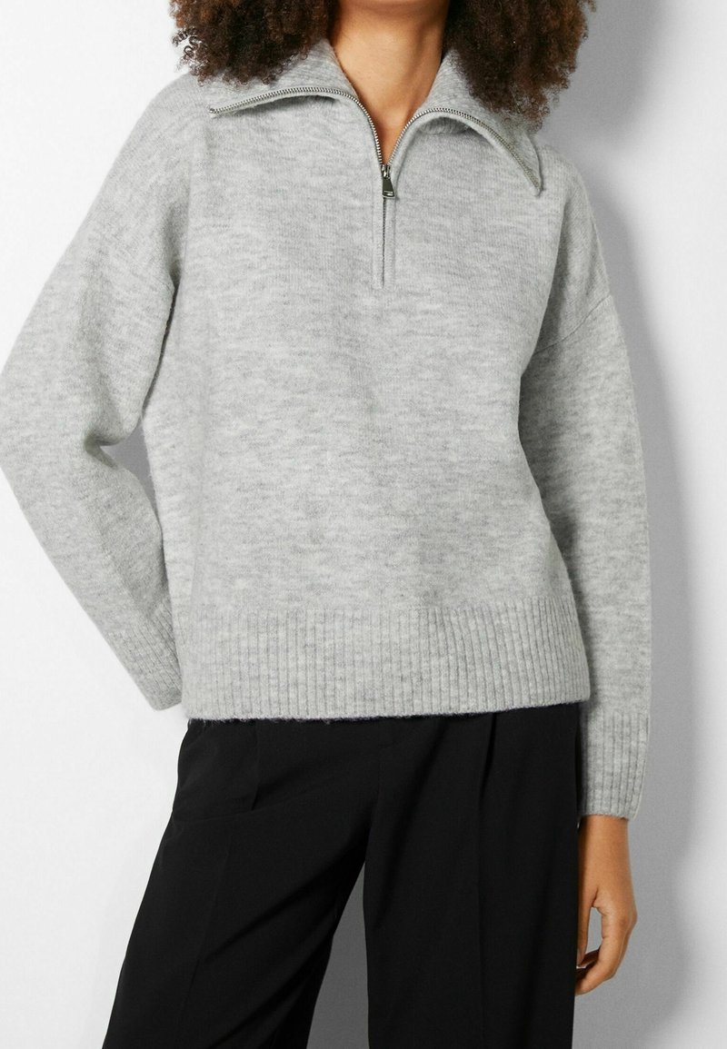 Suéter de punto en gris claro, con un cuello de media cremallera, dobladillo acanalado y mangas anchas, fabricado en un tejido suave y texturizado.