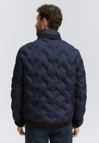 Man i marinblå vadderad jacka med svart krage och manschetter, ståendes vänd bort från kameran mot en enfärgad bakgrund.