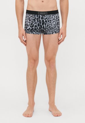 Mann in schwarzen und grauen Leopardmuster-Boxershorts mit Versace-Logo am Bund, barfuß vor einem schlichten Hintergrund stehend.