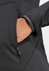 Detalj crne softshell jakne prikazuje ruku koja se pruža prema zatvorenoj pregradi, s glatkom teksturom i elegantnim dizajnom.