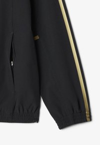 Zwarte jacksleeve met gouden verticale streep, elastische manchetten en gedeeltelijk zichtbare ritssluiting op een witte achtergrond.