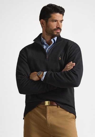 Maglione nero in pile con mezza cerniera, caratterizzato da un piccolo logo sul petto. Indossato sopra una camicia azzurro chiaro con collo e pantaloni beige.