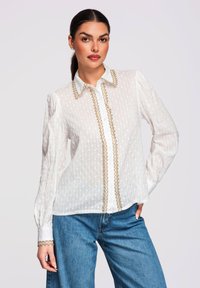 Femme portant un chemisier blanc texturé avec des bordures dorées et un jean bleu, debout avec une main sur la hanche devant un fond uni.