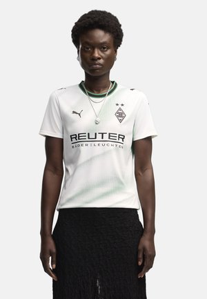 Persona con el pelo corto y rizado que lleva una camiseta blanca de fútbol del Borussia Mönchengladbach con logotipos de patrocinadores y una falda negra.