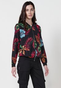 Floralna bomber jakna s intenzivnim uzorcima crvenih, plavih i zelenih listova na crnoj pozadini, prednjim zatvaračem i rebrastim manžetama.