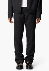 Zwarte broek van zachte stof met een ontspannen pasvorm, voorzien van een gladde textuur en twee zijzakken. Draagt zwarte geprinte schoenen.