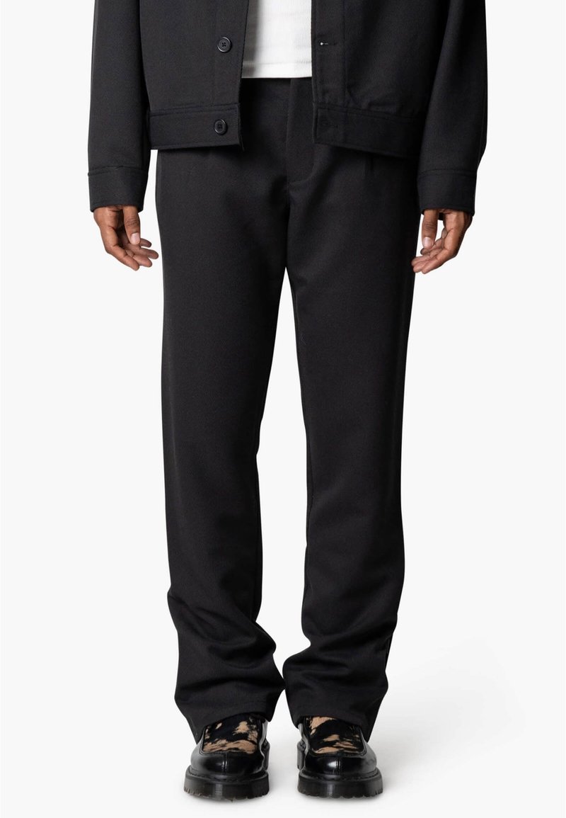 Zwarte broek van zachte stof met een ontspannen pasvorm, voorzien van een gladde textuur en twee zijzakken. Draagt zwarte geprinte schoenen.