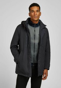 Parka imperméable marine avec une capuche amovible, fermeture éclair à l'avant et poches latérales, superposée à un pull zippé gris.