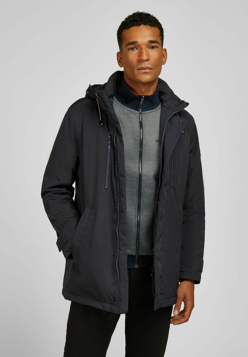 Parka imperméable marine avec une capuche amovible, fermeture éclair à l'avant et poches latérales, superposée à un pull zippé gris.