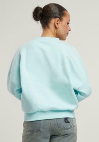 Felpa azzurra chiara con colletto a girocollo, vestibilità over size e orlo a costine. Indossata con jeans grigi leggermente sbiaditi con un'accentuazione sulla tasca posteriore.