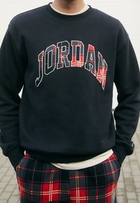 Person som bär en svart sweatshirt med "JORDAN" i röda rutiga bokstäver och matchande röda rutiga byxor, stående utomhus med händerna i fickorna.