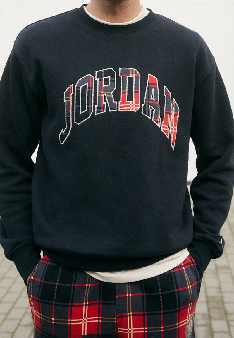Person som bär en svart sweatshirt med "JORDAN" i röda rutiga bokstäver och matchande röda rutiga byxor, stående utomhus med händerna i fickorna.