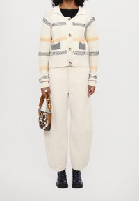 Sweter w kolorze kremowym z szarymi, pomarańczowymi i czarnymi paskami. Posiada duże guziki i dwie przednie kieszenie. Połączony z kremowymi spodniami typu wide-leg i wzorzystą torebką.