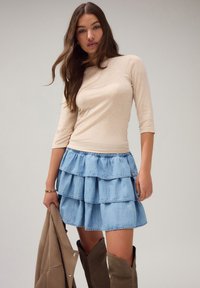 Kobieta w beżowej bluzce z długim rękawem, niebieskiej, warstwowej spódnicy dżinsowej, trzymająca beżową kurtkę i brązowe kozaki za kolano na jednolitym tle.