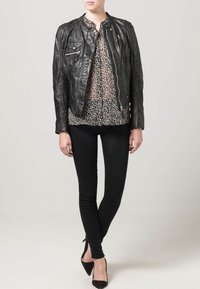 Schwarze Lederjacke mit tailliertem Design, ausgestattet mit Reißverschlusstaschen. Kombiniert mit einer schwarz-weiß gemusterten Bluse, schwarzen Skinny-Jeans und schwarzen High Heels.