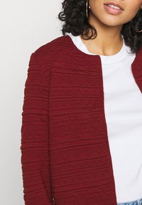 Cárdigan burdeos texturizado con patrones horizontales, cuello redondo y diseño de apertura frontal, llevado sobre una camisa blanca.