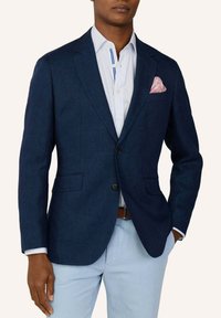 Mann trägt einen marineblauen Blazer mit einem rosa Einstecktuch, weiß gestreiftes Hemd, hellblaue Hose und braunen Gürtel, die Hand in der Tasche.
