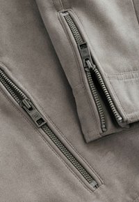 Tela de ante gris con dos cremalleras metálicas visibles en las mangas de la chaqueta, con textura suave y detalles de costura en contraste.